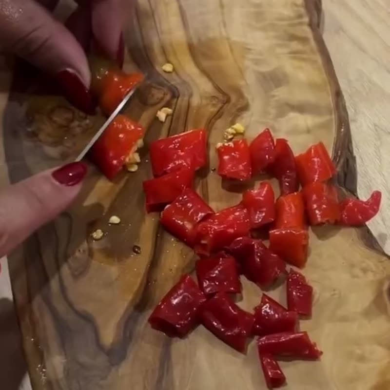 Des mains découpent des poivrons rouges sur une planche en bois d'olivier